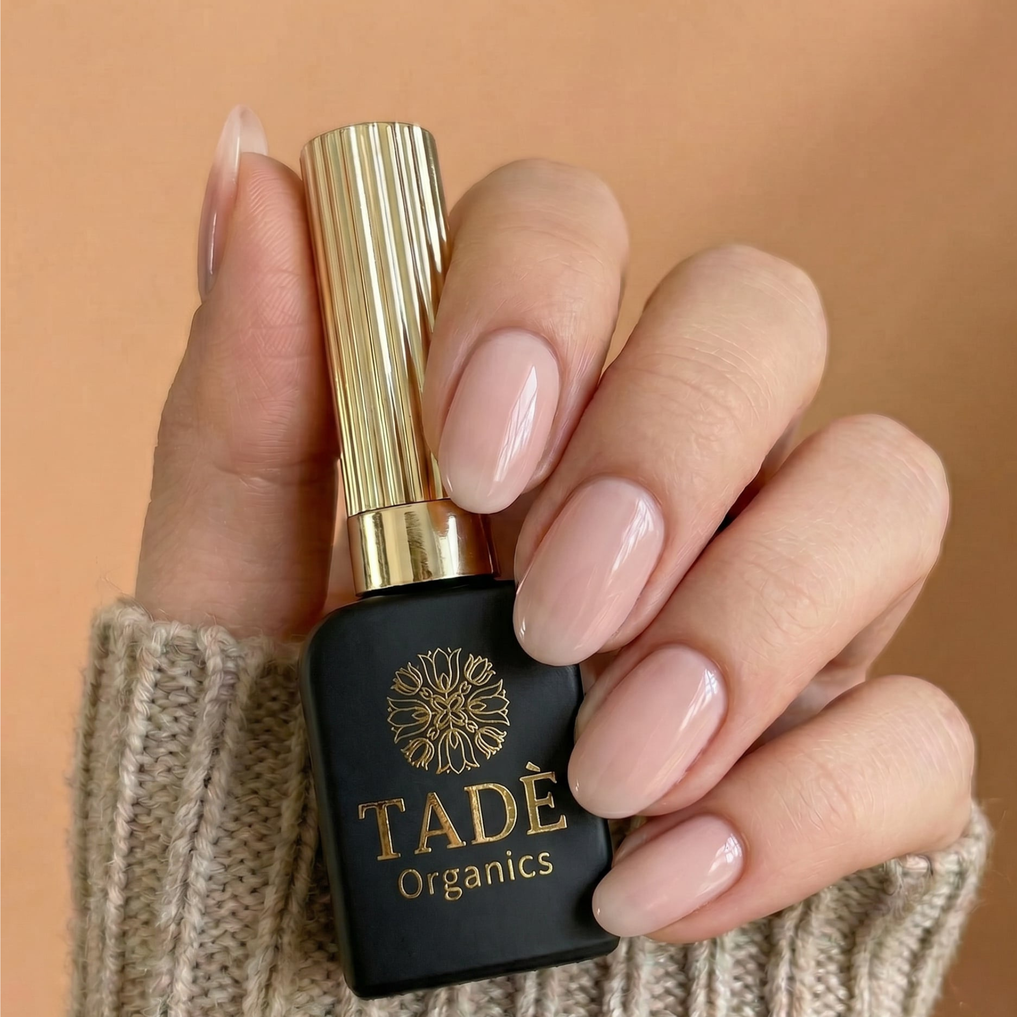 Hand met glanzende nude nagels die een zwart met gouden Tadè Organics nagellakflesje vasthoudt